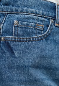 Κοντινό πλάνο μπλε τζιν με μπροστινή τσέπη που έχει πορτοκαλί ραφές και μεταλλικά καρφιά στις γωνίες της τσέπης.
