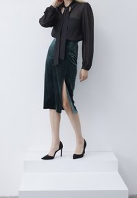 Blusa nera trasparente a maniche lunghe con colletto a fiocco e una gonna midi in velluto verde scuro con spacco laterale, abbinata a scarpe con tacco alte nere.