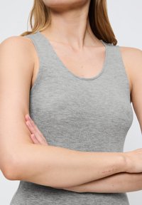 Szary bezrękawnik wykonany z miękkiej bawełny, z okrągłym dekoltem i gładką teksturą, z krzyżującymi się rękami i małą blizną widoczną na ramieniu.