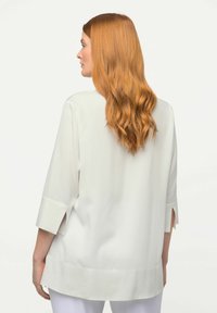 Weiße Bluse mit Dreiviertelärmel, offenen Seitenschlitzen und geradem Saum. Glatter Stoff mit minimalistischem Design.