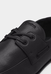 Μαύρο δερμάτινο παπούτσι με κορδόνια, με υφή και ραμμένα στοιχεία, καθώς και στρογγυλές θηλιές, με κοντινή λήψη από το μπροστινό και το πλάι.