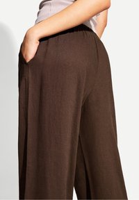 Brązowe, luźne spodnie z elastycznym pasem i kieszeniami po bokach; teksturowana tkanina nadaje głębi całemu projektowi.