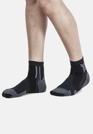 Jambes portant des chaussettes de sport noires et grises aux chevilles avec des motifs tricotés texturés et des logos sur les zones du talon et des orteils.