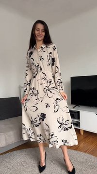 Vestido longo bege com padrões florais pretos, gola com botões, mangas compridas e saia rodada. Usado com sapatos pretos de salto alto.