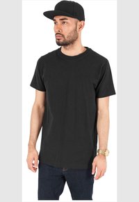 T-shirt en coton noir à manches courtes et col rond, associée à une casquette noire et un jeans en denim foncé. Montre dorée au poignet.