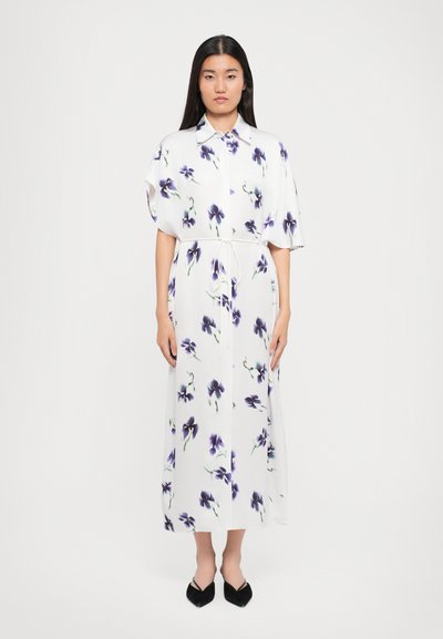Robe blanche à motifs floraux avec manches bouffantes, devant à boutons et détail à nouer à la taille. Assortie à des chaussures noires, au design plat.