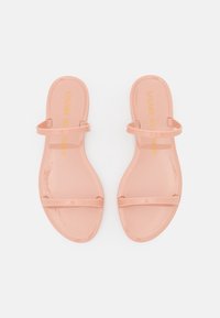 Sandales à talons plats en vernis rose avec deux fines lanières sur le dessus du pied. La semelle est plate et présente le nom de la marque en relief en jaune.