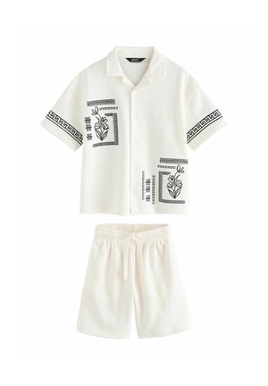 Dreng med krøllet hår iført hvidt kortærmet sæt med shorts med sorte geometriske og blomsteragtige mønstre, der står op ad en lys væg.