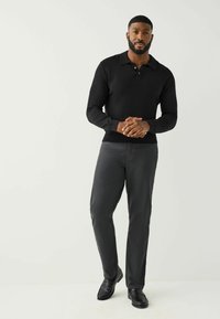 Juodas ilgomis rankovėmis megztinis su apykakle, keturiais mygtukais ir gofruotomis rankogaliais, derinamas su tamsiai pilkomis kelnėmis ir juodais batais.