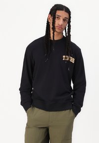 Zwarte sweatshirt met lange mouwen en geribde manchetten. Heeft een logo-patch in beige op de borst. Draagt met olijfgroene cargobroek.