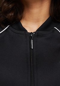 Svart zip-up jacka med hög krage, med en slät yta, vita detaljer längs axlarna och en metallisk dragkedja.