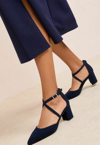 Tumesinised seemisnahast kontsakingad terava varbaotsaga, paksu kand, ning ümber pahkluu risti käiv rihm, millel on väike metallist pandla detail.