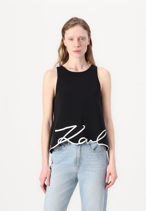 Top corto negro sin mangas hecho de una tela suave, con un detalle en cursiva blanca que dice "Karl" en el dobladillo. Combinado con jeans de color azul claro.