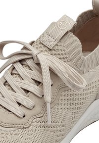 Gros plan sur une basket beige en tricot avec des lacets épais et arrondis noués en un nœud et une languette en tissu à l'arrière de la tige.