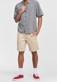 Camicia a maniche corte grigia con bottoni e pattern multicolor, abbinata a pantaloni corti beige e sneakers rosse, che mostrano un fit casual e rilassato.