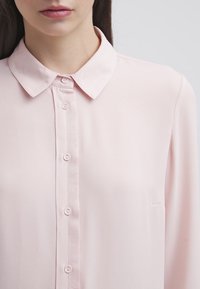 Blusa lunga a maniche lunghe color rosa chiaro, con apertura a bottoni frontale, colletto e taschino sul petto. Realizzata in tessuto morbido e liscio.