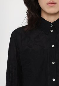 Zwarte, sheer button-up blouse met een textuurrijk bloemenpatroon, voorzien van een klassieke kraag en parelmoerachtige knopen langs de voorkant.