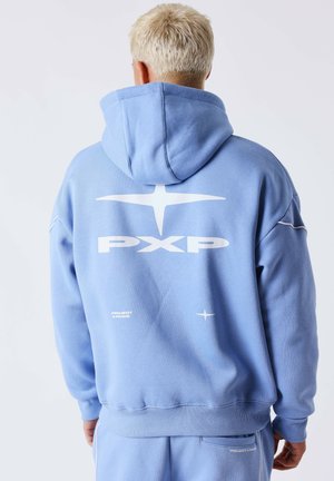 Persona con capelli biondi corti che indossa una felpa con cappuccio e pantaloni azzurri chiaro con il logo bianco "PXP" e una stella sul retro della felpa.
