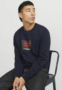 Marineblauwe katoenen sweatshirt met een zwart en rood grafisch logoontwerp op de voorkant. Lange mouwen en geribbelde manchetten. Draagt men met donkere jeans.