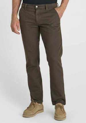 Man draagt donkerbruine rechte broek met beige casual schoenen, één hand in de zak, staand tegen een effen achtergrond.
