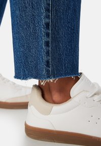 Sneaker bianco con dettagli beige, suola in gomma texturizzata, abbinato a jeans in denim blu con orlo sfilacciato e taglio dritto.