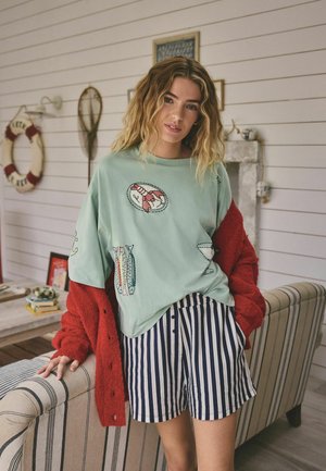 Young woman wearing a mint green top with seafood and drink embroidery, striped shorts, and a red cardigan, leaning on a striped sofa.