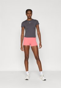 Ανδρικό εφαρμοστό γκρι μπλουζάκι Nike με κοντά μανίκια, σε συνδυασμό με ροζ αθλητικά σορτς. Το μοντέλο φοράει λευκά αθλητικά παπούτσια και κάλτσες με το λογότυπο της Nike.