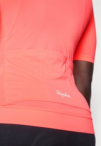 Trumpas rankovėmis dviratininko džemperis ryškiai koralinės spalvos, su tekstūruota raštu, užpakalini pocket'ą ir baltu "Rapha." logotipu arti krašto.