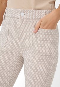 Lichtbeige patroonbroek met geometrische vormen, voorzien van een zak aan de voorkant en een knoopsluiting in de taille. Geweven stofontwerp.