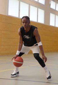 Basketballspiller i en svart ermeløs trøye og hvite shorts, som dribler en basketball på et gymsgulv, iført svart og hvite joggesko.