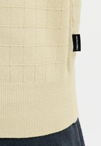 Ourlet de pull beige en maille avec poignets côtelés, motif à carreaux dessus et étiquette noire de la marque Jack & Jones sur la couture latérale.