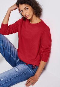 Czerwona bluza z długim rękawem i prążkowanym dekoltem, zestawiona z niebieskimi rurkami z przetarciami. Jeansy mają wyblakły wzór i widoczne szwy.