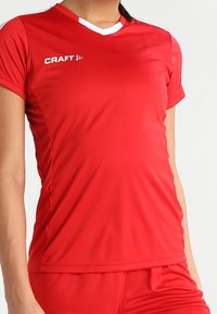 Rød syntetisk sportstrøje med korte ærmer, V-udskæring, hvide detaljer og meshnätter i siden. Har logo på brystet.