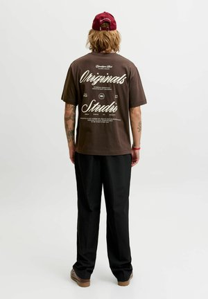 Person mit Tattoos, die ein braunes Grafik-T-Shirt mit der Aufschrift "Originals Studio,", schwarze Hose, rote Kappe und braune Schuhe trägt, von hinten auf weißem Hintergrund.
