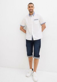 Camicia bianca a maniche corte con colletto e polsini a scacchi, abbinata a pantaloni cargo blu scuro arrotolati e sneaker bianche.
