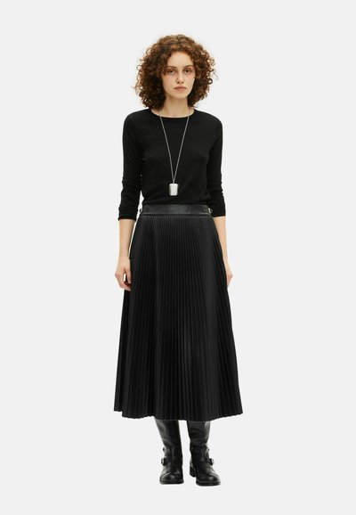 Γυναίκα με σγουρά μαλλιά, φοράει μαύρο μπλουζάκι μακρύ μανίκι, μαύρη πλισέ midi φούστα, μαύρες μπότες και μια μακριά ασημένια κρεμαστή αλυσίδα.