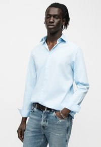 Νέος άντρας με σκούρα πλεξούδες φοράει ανοιχτό μπλε πουκάμισο με κουμπιά και ξεθωριασμένο μπλε τζιν παντελόνι, στέκεται με το ένα χέρι στην τσέπη μπροστά από απλό φόντο.