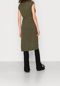 Kvinna som står med ryggen vänd, iklädd en olivgrön stickad ärmlös klänning över svarta utsvängda byxor och svarta platåsandaler.