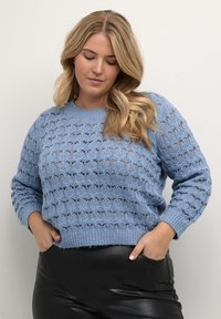 Jasnoniebieski sweter z dzianiny, charakteryzujący się teksturowanym, otwartym wzorem koronkowym, okrągłym dekoltem i ściągaczami, w zestawie z czarnymi skórzanymi spodniami.
