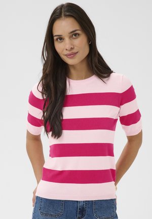 Kvinde med langt mørkt hår iført en kortærmet pink og mørk pink stribet sweater og blå jeans, stående med hænderne bag ryggen.