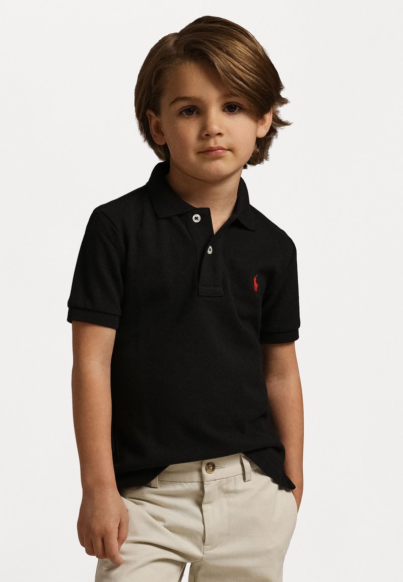 Schwarzes Polohemd mit zwei Knöpfen und kurzen Ärmeln, mit einem kleinen roten Logo auf der Brust, getragen mit hellbeigen Hosen.