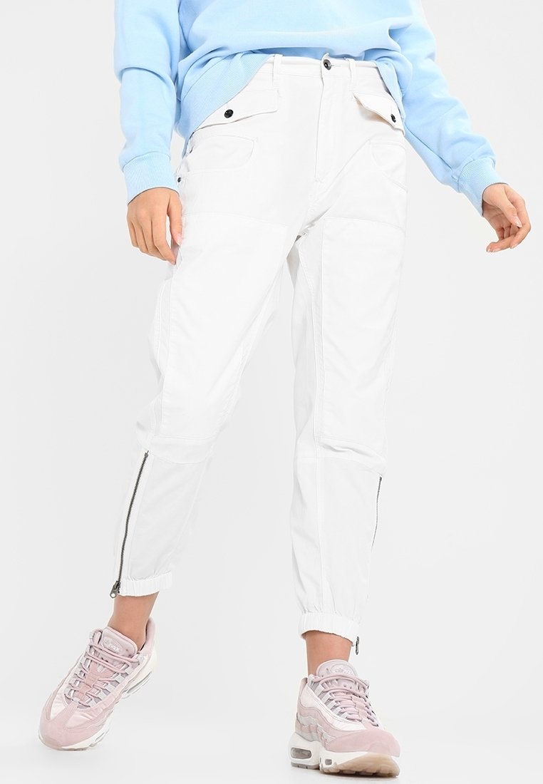 Personne portant un pantalon cargo blanc avec des fermetures éclair aux jambes et un pull bleu clair, associé à des baskets roses et blanches.