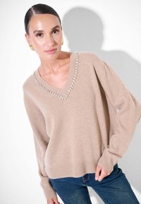 Maglione beige a v con orlo e maniche a costine, impreziosito da strass attorno al colletto. Indossato con jeans in denim blu.