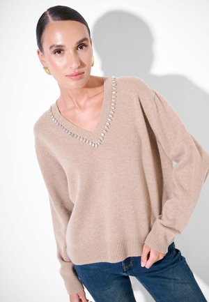 Maglione beige a v con orlo e maniche a costine, impreziosito da strass attorno al colletto. Indossato con jeans in denim blu.