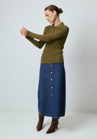 Oliivinvihreä ribattuja pitkähihainen polo-paita yhdistettynä tummansinisään denim-hameeseen, jossa on kuparinvärisiä nappeja. Jalassa on ruskeat kärjelliset nilkkurit.