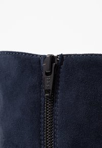 Bottine en daim bleu marine avec une fermeture visible par une fermeture éclair noire YKK et des coutures plates le long de l'ouverture pour la fermeture et le détail.