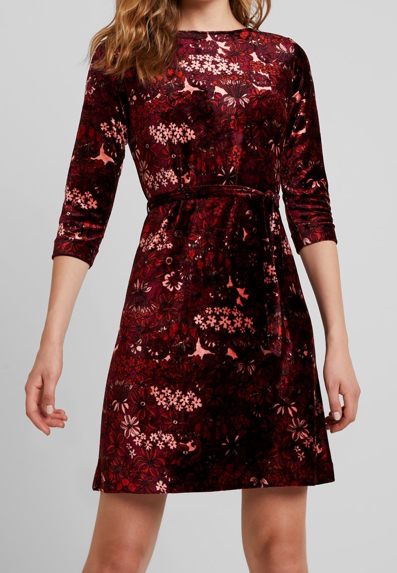 Femme portant une robe en velours bordeaux mi-longue à manches longues, avec un motif floral et une ceinture nouée à la taille.