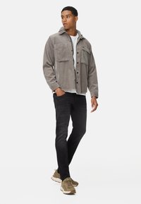 Grau gestreifte Cordjacke mit zwei Fronttaschen und Kapuze, kombiniert mit schwarzen Slim-Fit-Jeans und olivgrünen Sneakers, stehend vor einem weißen Hintergrund.