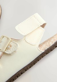 Sandales beige clair avec brides réglables en Velcro, dessus en cuir synthétique texturé, semelle intérieure en liège et semelle extérieure en caoutchouc marron.