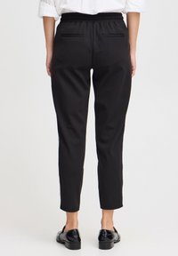 Mustat, joustavavyötäröiset housut, joissa on kaksi takataskua ja kapea istuvuus. Sileä kangas sivusaumoilla. Yhdistetty kiiltäviin mustiin kenkiin.